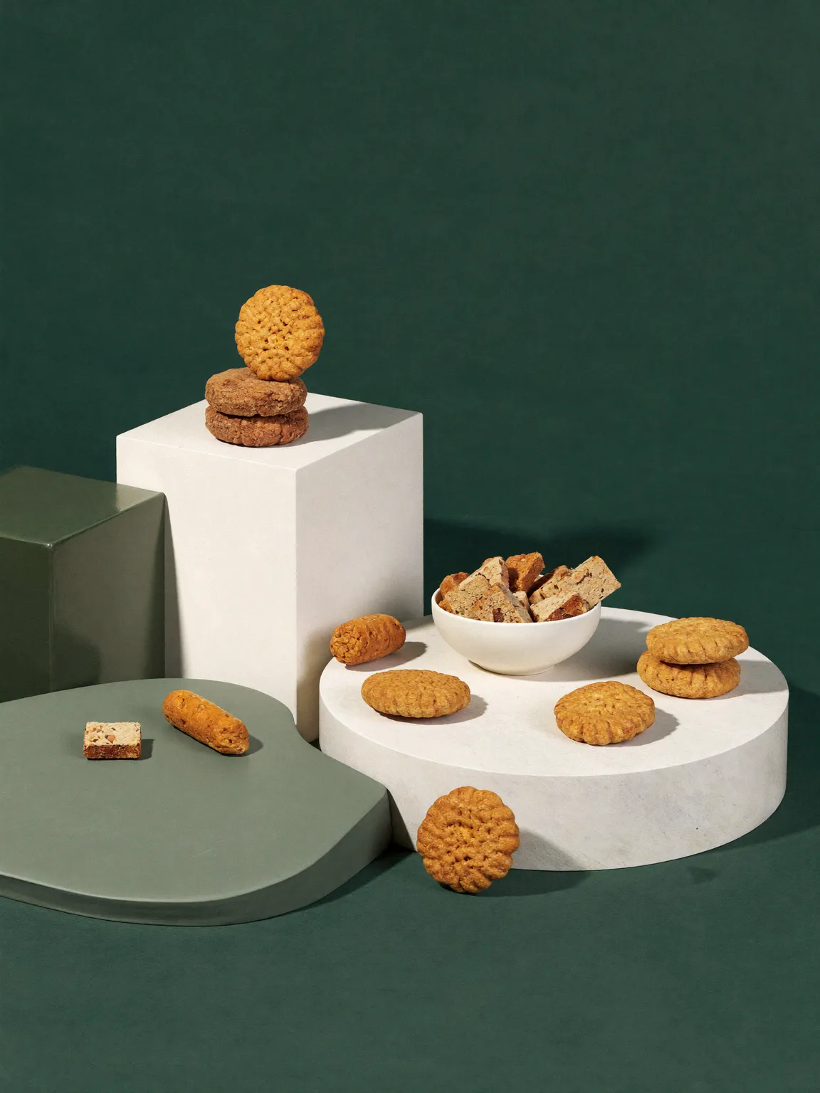 Dog biscuits and treats arranged on pale plinths against a deep green backdrop.